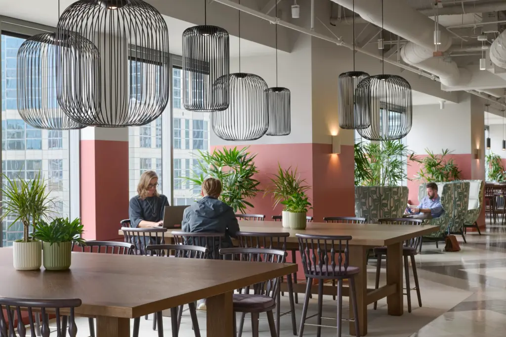 Modern office lounge with large windows, wooden tables, potted plants, and stylish black wire pendant lights. People are working and conversing in the open, well-lit space.